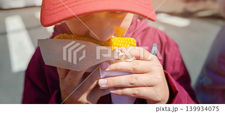 Child wearing red cap enjoys eating corn on the cob while holding it with both hands, outdoor setting with blurred background elements visible Child wearing red cap enjoys eating corn on the cob while holding it with both hands, outdoor setting with blurred background elements visible 139934075