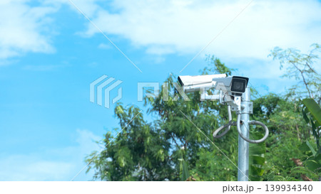 White outdoor security CCTV camera mounted on a metal pole against a vibrant blue sky with light clouds. Lush green tree branches and foliage are visible in the background frame. 139934340