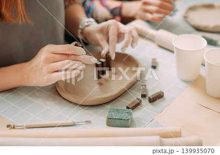 close-up female hands make a clay plate, align the shape with special tools in the workshop close-up female hands make a clay plate, align the shape with special tools in the workshop 139935070