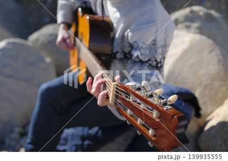 A performer of songs with a guitar. A woman plays the guitar, close-up, focus on the strings and neck of the guitar. The musician is sitting on large rocks 139935555