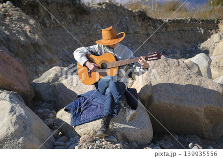 A female cowboy plays guitar sitting on a large rock surrounded by other boulders. A musician learns to play the guitar on a sunny day on a pile of stones. The concept of freedom and adventure 139935556
