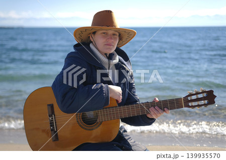 A lonely woman in a cowboy hat is sitting on the shore of a lake with a guitar in her hands. The beach is quiet and peaceful, and the guitarist seems to be enjoying playing the guitar 139935570