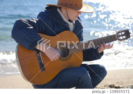 A guitarist on the beach in the warm rays of the sun. A woman in a cowboy hat plays guitar on the ocean. The musician is wearing a blue jacket A guitarist on the beach in the warm rays of the sun. A woman in a cowboy hat plays guitar on the ocean. The musician is wearing a blue jacket 139935571