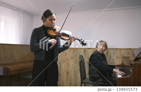 An Asian teenage girl plays the violin to the accompaniment of a piano. An elderly Asian woman conducts a violin lesson while sitting on a chair and playing the piano. Music school An Asian teenage girl plays the violin to the accompaniment of a piano. An elderly Asian woman conducts a violin lesson while sitting on a chair and playing the piano. Music school 139935573