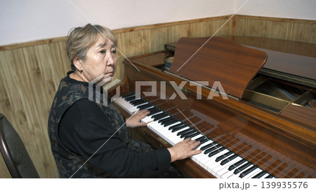 An elderly Asian woman plays the piano in a music school classroom. She's focused on her game. Piano lessons. An old pianist at work. A musician An elderly Asian woman plays the piano in a music school classroom. She's focused on her game. Piano lessons. An old pianist at work. A musician 139935576