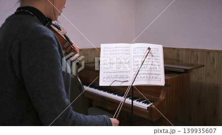 A young Asian woman is learning to play the violin in a room with a piano. She holds a music stand with music sheets in front of her. Music school. A student in a violin lesson A young Asian woman is learning to play the violin in a room with a piano. She holds a music stand with music sheets in front of her. Music school. A student in a violin lesson 139935607