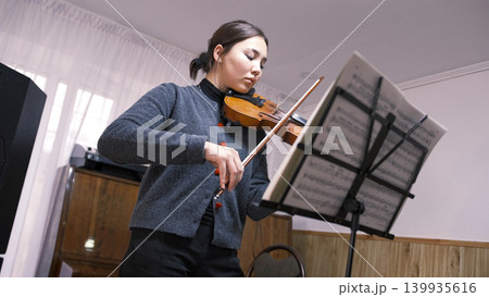 An Asian teenage student is learning to play the violin. A young woman looks at the sheet music and plays the violin at a music school. The musician is focused on the rehearsal 139935616