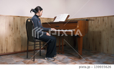 A young Asian student is learning to play the piano. Music school. A woman is sitting at the piano with a music book in front of her. She plays the piano and is focused on music 139935626