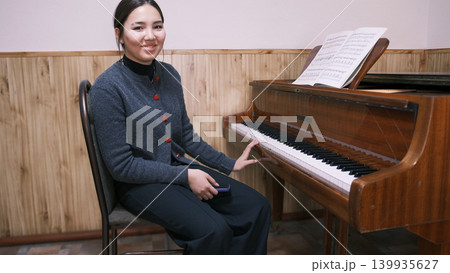 Portrait of a happy Asian student at the piano with sheet music. A young woman is sitting in front of a piano in a music school classroom. The teenage musician is smiling 139935627