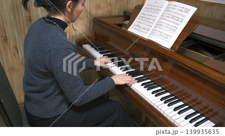 A young Asian schoolgirl is learning to play the piano by sheet music. A woman is playing the piano with sheet music spread out in front of her A young Asian schoolgirl is learning to play the piano by sheet music. A woman is playing the piano with sheet music spread out in front of her 139935635