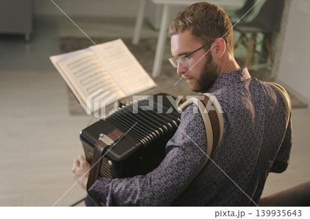 A bearded guy with glasses is playing the accordion, there are notes next to him. The musician holds a small accordion in his hands and learns a new melody. The accordion player 139935643