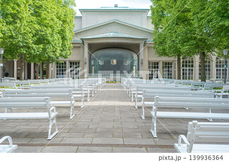 Symmetric view of empty white wooden benches arranged for an outdoor concert in front of the music pavilion. Green trees and pavement in a quiet spa resort 139936364