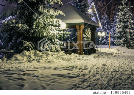 Gazebo, wooden house and Christmas trees in the snow at night. A trodden door leads to the gazebo. Lanterns and garlands illuminate the gazebo and the house. Winter resort in the mountains Gazebo, wooden house and Christmas trees in the snow at night. A trodden door leads to the gazebo. Lanterns and garlands illuminate the gazebo and the house. Winter resort in the mountains 139936492