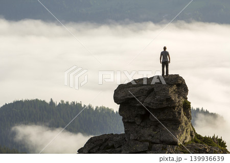 Silhouette of athletic climber tourist on high rocky formation on mountain valley filled with white puffy clouds and fog and covered with evergreen forest mountain slopes under clear sky background. Silhouette of athletic climber tourist on high rocky formation on mountain valley filled with white puffy clouds and fog and covered with evergreen forest mountain slopes under clear sky background. 139936639