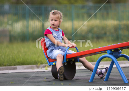 Small cute young blond child girl sits moody, angry and unsatisfied on see- saw swing on warm sunny day on bright green blurred background. Joys and sorrows of childhood, children tantrums concept. 139936657