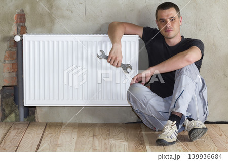 Young handsome plumber sitting on floor with wrench in hand near successfully installed heating radiator in empty room of a newly built apartment or house. Construction, maintenance and repair concept Young handsome plumber sitting on floor with wrench in hand near successfully installed heating radiator in empty room of a newly built apartment or house. Construction, maintenance and repair concept 139936684