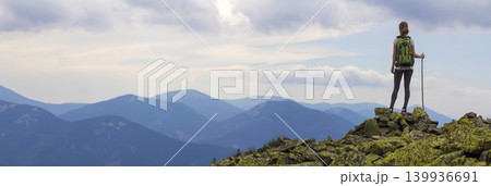 Back view of young slim backpacker tourist girl with stick standing on rocky top against bright blue morning sky enjoying foggy mountain range panorama. Tourism, traveling and climbing concept. 139936691