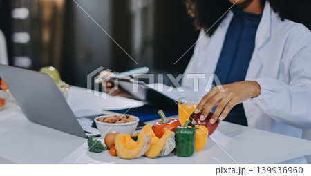 Young nutritionist showing bell peppers during an online video call consulting about healthy eating, sitting at home at a table with vegetables, laptop and measuring tape Young nutritionist showing bell peppers during an online video call consulting about healthy eating, sitting at home at a table with vegetables, laptop and measuring tape 139936960
