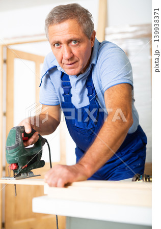 Experienced male worker cuts board with an electric jigsaw in room being renovated Experienced male worker cuts board with an electric jigsaw in room being renovated 139938371