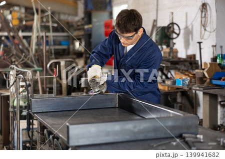 Young worker uses an angle grinder while working at mechanical engineering plant Young worker uses an angle grinder while working at mechanical engineering plant 139941682