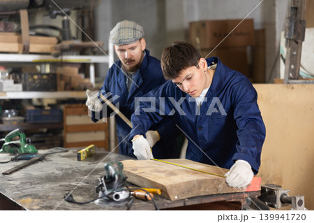 Carpenter measures the length of board with tape measure. Foreman prompts him 139941720