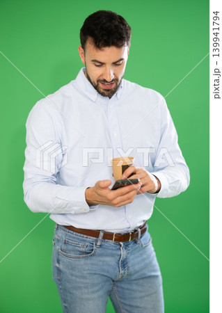 Adult man drinking coffee and scrolling through her smartphone screen against green screen background. Chroma key compositing 139941794