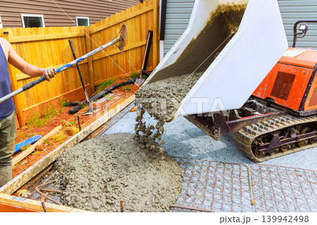 Worker uses tracked dumper mixer to pour concrete into framework for patio in backyard area. 139942498