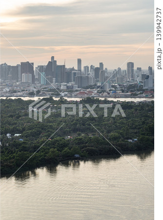 An emerald canopy covers the Bang Kachao peninsula, a lush sanctuary against the modern world. In the foreground, the Chao Phraya River ripples softly under a muted golden sky. Across the water, the 139942737
