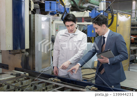 製造業の工場現場で作業着の技術者と営業が機械を見ながら見積もりや修理、メンテナンスの金額を前向き 製造業の工場現場で作業着の技術者と営業が機械を見ながら見積もりや修理、メンテナンスの金額を前向き 139944231