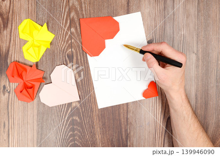 male hand writes with a pen on a white sheet of paper with colorful origami hearts male hand writes with a pen on a white sheet of paper with colorful origami hearts 139946090