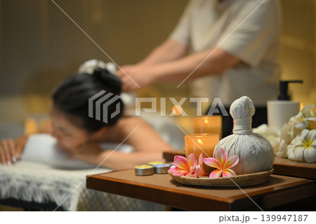 Focus on spa equipment and relaxing candlelight woman receiving traditional herbal compress massage in a serene spa 139947187