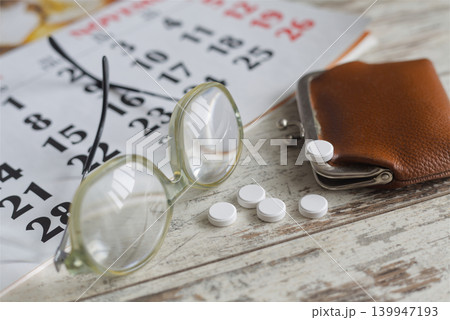 Conceptual close-up of reading glasses, white pills, and an old wallet on a calendar background on a wooden table. Conceptual close-up of reading glasses, white pills, and an old wallet on a calendar background on a wooden table. 139947193