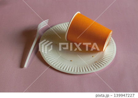 Orange disposable cup lying on a disposable plate with crumbs after food, next to a disposable fork on a pink background. Orange disposable cup lying on a disposable plate with crumbs after food, next to a disposable fork on a pink background. 139947227
