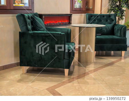 Elegant emerald velvet armchairs near electric fireplace in luxury lobby Elegant emerald velvet armchairs near electric fireplace in luxury lobby 139950272