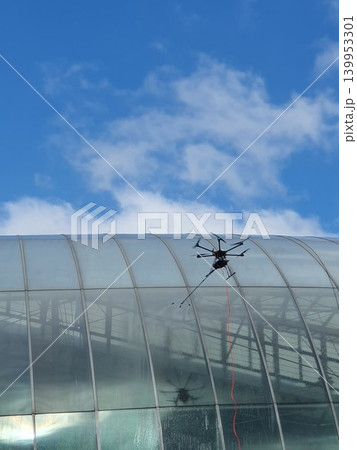A tethered inspection drone hovers beside a curved glass roof of a modern building, conducting maintenance and window cleaning under a bright blue sky with scattered clouds, urban drone technology. A tethered inspection drone hovers beside a curved glass roof of a modern building, conducting maintenance and window cleaning under a bright blue sky with scattered clouds, urban drone technology. 139953301
