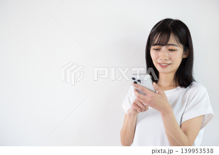 白い背景の前でスマートフォンを持ちながら微笑む日本人の若い女性 139953538