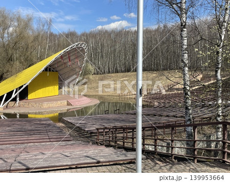 Outdoor amphitheater with empty wooden seating surrounded by trees under a blue sky 139953664