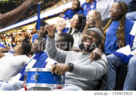 Black football supporter smiling and beating drum in blue stadium stands, leading energetic crowd chants during live soccer match. Soccer ultras creating loud stadium chants with pride. 139953925