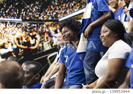 Diverse blue team football supporters chanting in packed stadium stands during live soccer match, fan scarves and excited crowd atmosphere. Father, mother and son sharing family fun with laughter. Diverse blue team football supporters chanting in packed stadium stands during live soccer match, fan scarves and excited crowd atmosphere. Father, mother and son sharing family fun with laughter. 139953944