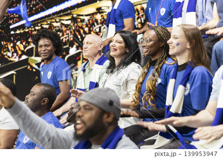 Diverse blue team women supporters cheering loudly with raised arms in packed stadium stands during exciting live soccer match. Team spirit and celebration inside the stadium terrace. Diverse blue team women supporters cheering loudly with raised arms in packed stadium stands during exciting live soccer match. Team spirit and celebration inside the stadium terrace. 139953947