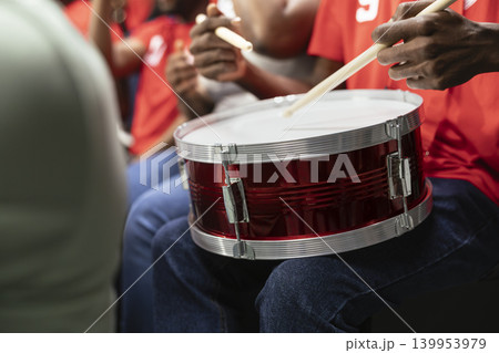 Close-up of red supporter drum on football fan lap during stadium chant, with cheering crowd and scarves in background. Organized fans cheering and creating powerful fan chants. Close-up of red supporter drum on football fan lap during stadium chant, with cheering crowd and scarves in background. Organized fans cheering and creating powerful fan chants. 139953979