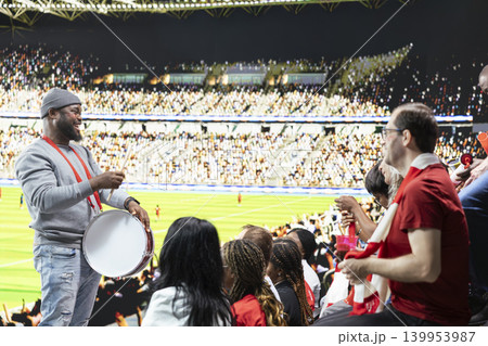 Energetic fan leader standing in the terrace section with drum, organizing chanting fans during live football match. Diverse football ultras create loud noise with percussion, stadium crowd. Energetic fan leader standing in the terrace section with drum, organizing chanting fans during live football match. Diverse football ultras create loud noise with percussion, stadium crowd. 139953987