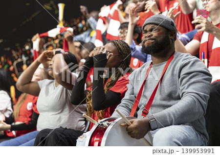 Black football supporter with drum closing eyes and holding breath in stadium stands during tense live soccer match moment, Diverse fans sitting in silence and disbelief, losing team. Black football supporter with drum closing eyes and holding breath in stadium stands during tense live soccer match moment, Diverse fans sitting in silence and disbelief, losing team. 139953991