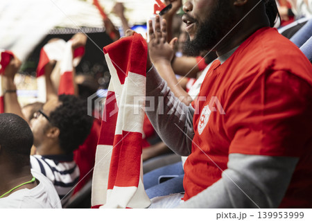 Organized fan club waving scarves celebrating in the terrace stands, thrilling football match in a crowded stadium. Active fans create synchronized fan chants, match excitement and celebration. 139953999
