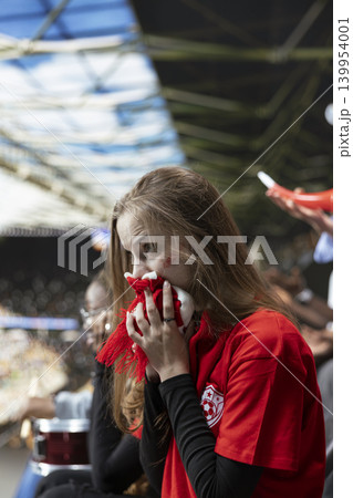 Close portrait of anxious young female football supporter holding red scarf to mouth during dramatic live match in stadium crowd. Nervous speechless woman with face paint, equality in sports. 139954001