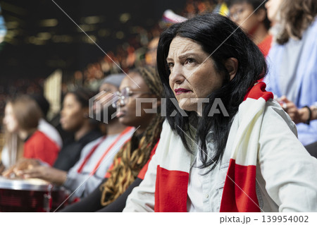 Close up of mature female football fan with red scarf watching match suspense in stadium stands, worried expression and crowd behind. Anxious supporters watching decisive final minutes in silence. 139954002