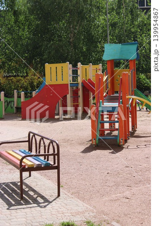 Colorful playground features slides and climbing frames next to a striped bench in a sandy area with green trees and grass in the background 139954867