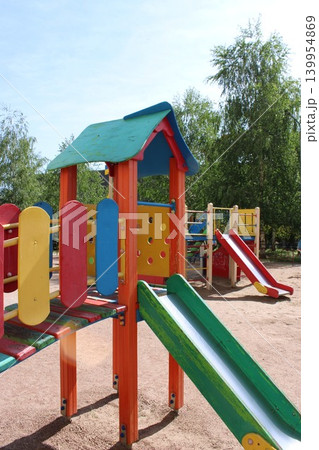 Colorful children's playground equipment featuring slides, climbing structures, and play panels in a sunny outdoor setting surrounded by trees and blue sky 139954869