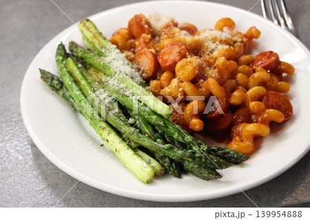 Cooked pasta with tomato sauce and sausage served with grilled asparagus sprinkled with cheese on a white plate against a gray background 139954888