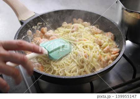 Cooking shrimp pasta in a black frying pan with a green spatula, steam rising, and a pot of boiling water visible in the background on a modern kitchen stove Cooking shrimp pasta in a black frying pan with a green spatula, steam rising, and a pot of boiling water visible in the background on a modern kitchen stove 139955147